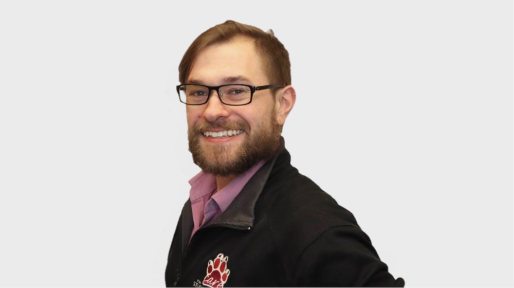 Professional headshot of a smiling man with glasses and a beard on a light background
